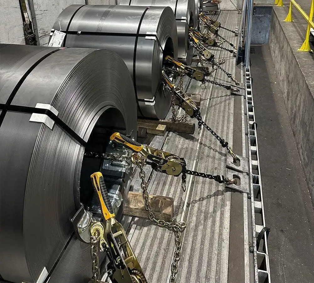 Steel coils loaded eyes-crosswise on a flatbed trailer, secured with grade 70 chain and lever binders through the eye of each coil, sitting on coil racks with wood dunnage